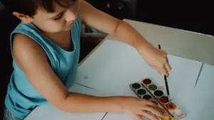 Mały chłopiec malujący akwarelami na białym arkuszu papieru w jasnym pomieszczeniu.