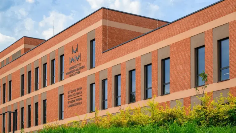 Nowoczesny budynek z czerwonej cegły z logo Uniwersytetu Warmińsko-Mazurskiego na słonecznym tle nieba.
