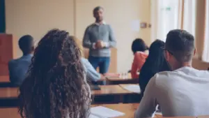 Studentki i studenci w auli uniwersyteckiej słuchający wykładu prowadzonego przez nauczyciela.