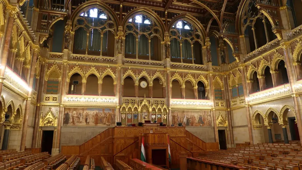 Wnętrze sali obrad parlamentu z bogatymi zdobieniami architektonicznymi i drewnianymi ławami poselskimi.