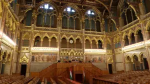 Wnętrze sali obrad parlamentu z bogatymi zdobieniami architektonicznymi i drewnianymi ławami poselskimi.